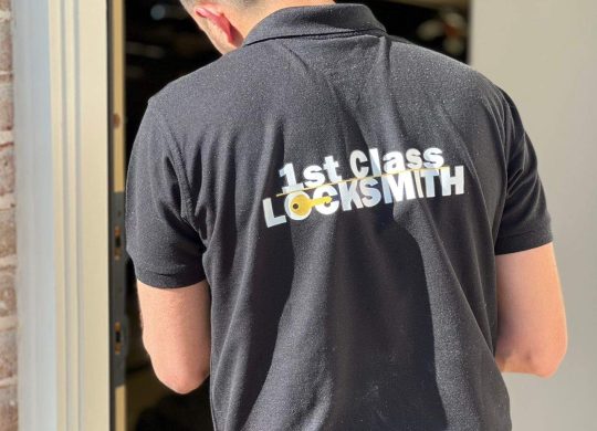 1st Class Locksmith technician working on a residential door lock, wearing a black uniform with yellow company branding in Fort Worth, TX